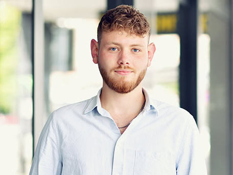 Ein junger Mann mit lockigem Haar und Bart lächelt in einem weißen Hemd.