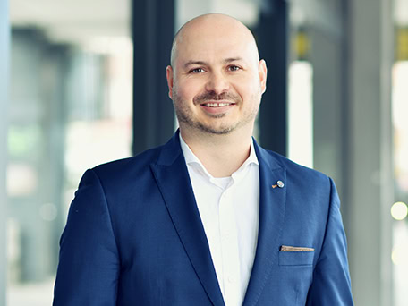 Ein lächelnder Mann im blauen Anzug mit Glatze vor unscharfem Hintergrund.