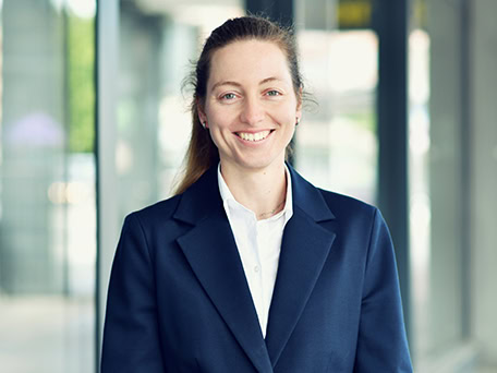 Eine lächelnde Frau in dunkelblauem Blazer vor einem modernen Gebäude.