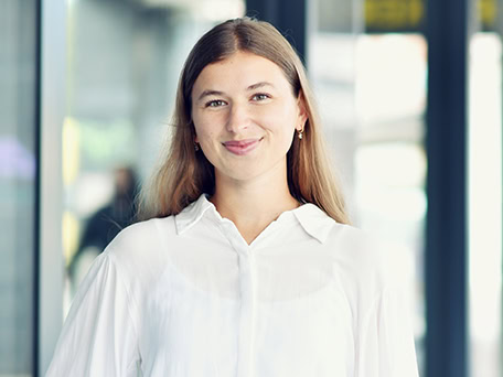 Junge Frau lächelt in weißer Bluse, lange Haare, vor unscharfem Hintergrund.