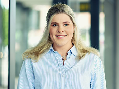 Eine lächelnde Frau mit blonder Kleidung und offenem Hemd steht vor einem verschwommenen Hintergrund.